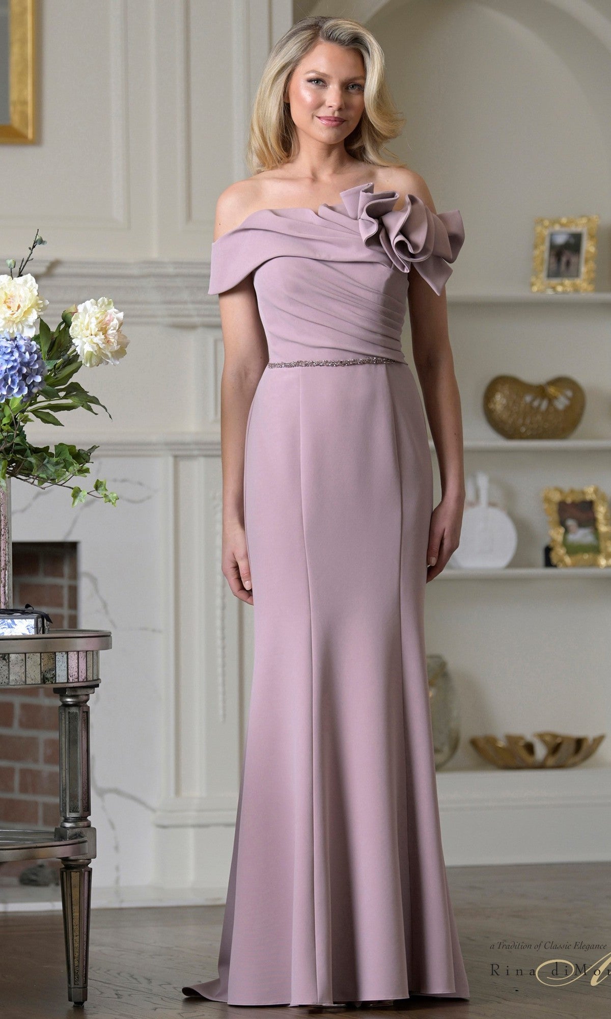 A woman in a Rina Di Montella RD3507 long formal dress in dusty rose, featuring an off-the-shoulder design and floral bodice detail, stands in a stylishly decorated living room.